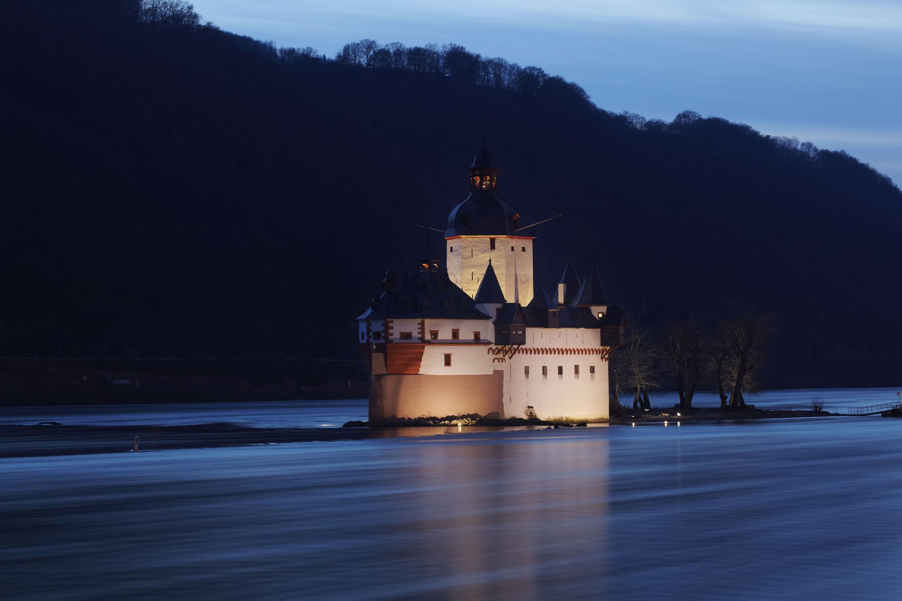 Die beleuchtete Burg Pfalzgrafenstein am Abend im Rhein, dahinter ein dunkler Hang