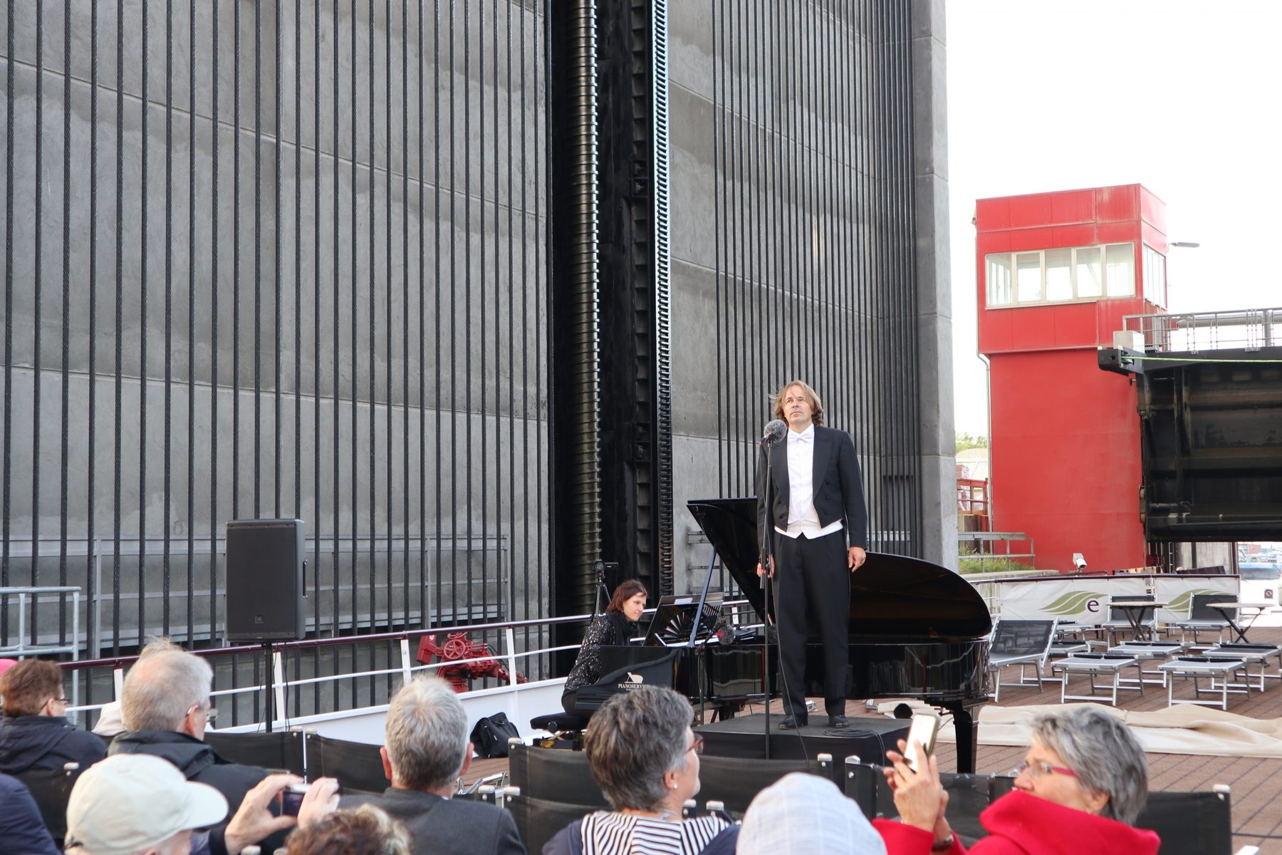 Ein Sänger am Flügel auf dem Schiffsdeck vor der hohen Wand des Hebewerks, daneben eine Pianistin, davor Zuhörer