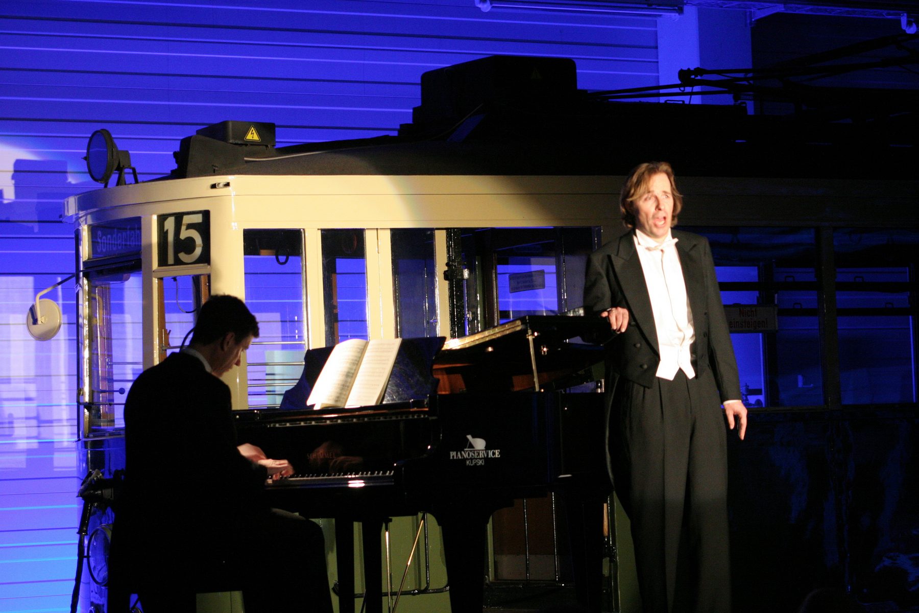 Der Bariton im Frack neben dem Flügel in blau beleuchteter Halle, davor eine historische Straßenbahn mit der Nummer 15 und ein Pianist
