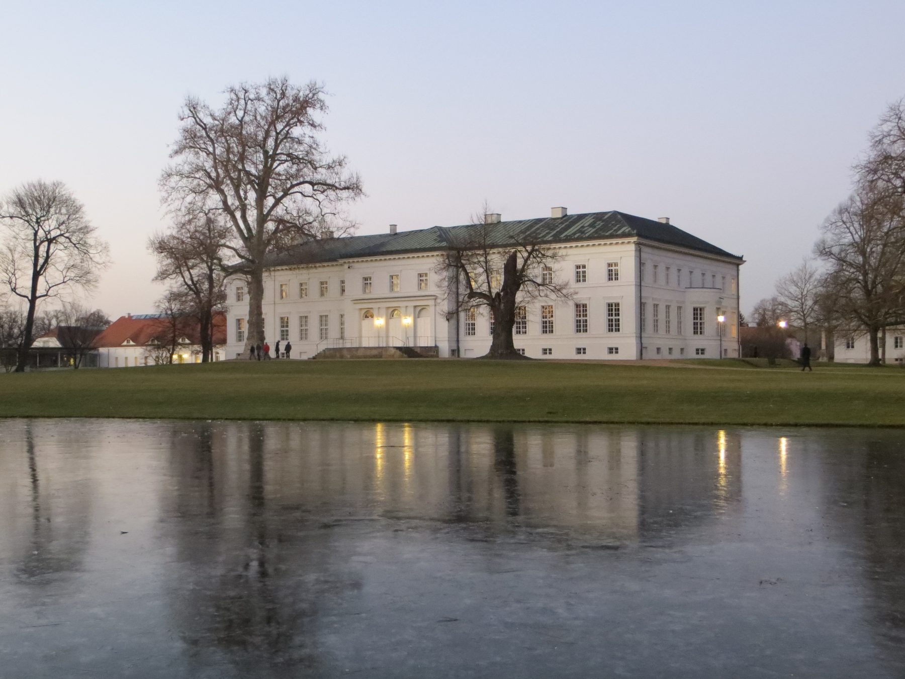 Schloss Neuhardenberg in der Dämmerung, davor der zugefrorene Teich, in dem sich die erleuchteten Fenster spiegeln