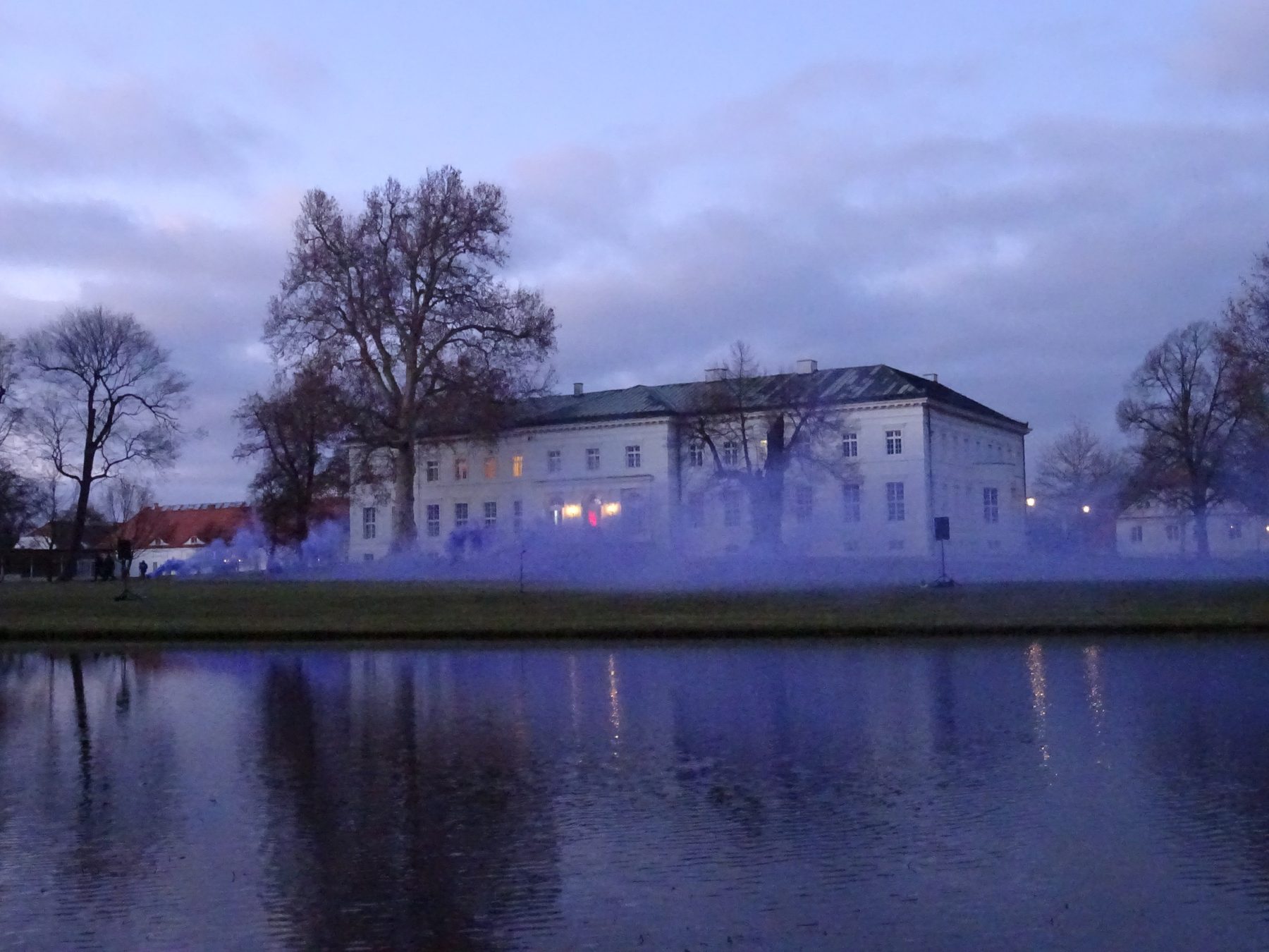 Schloss Neuhardenberg in der Dämmerung, davor das Wasser und blauer Nebel, in dem sich die Lichter spiegeln
