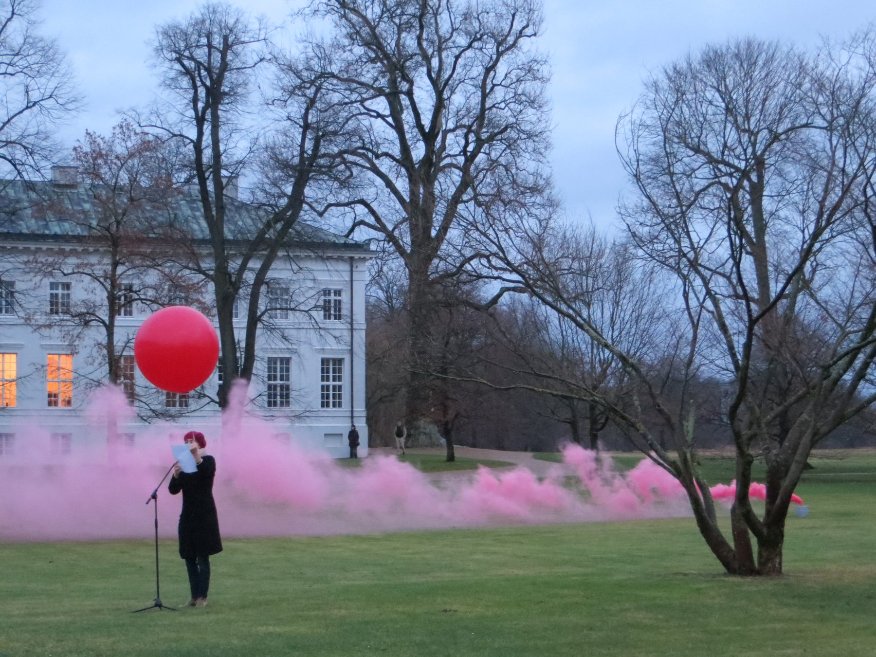 Vor dem Schloss steht eine Frau mit Mikrofon im rosa Rauch, über ihr schwebt ein roter Ballon